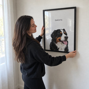 A woman in a black hoodie hangs a framed custom pet portrait on a white wall. The portrait features a black, white, and tan Bernese Mountain Dog with the name 'DAKOTA' printed at the top.